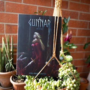 May include: A rectangular comic book titled Gunnar and the Mage by Valerio Pozzi. The cover features a blonde character in a red coat standing in a dark forest, holding a sword. The title is written in white stylized lettering at the top.