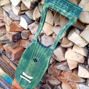 May include: A green wooden lyre with five strings, a sound hole, and a decorative design on the top. The lyre is resting on a pile of firewood.