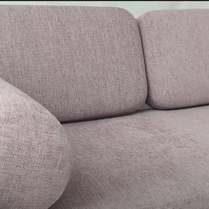 Puede incluir: Un sofá de color lila claro con un cojín grande y cilíndrico en el reposabrazos izquierdo. El sofá tiene una tapicería de tela capitoné y un marco de madera.