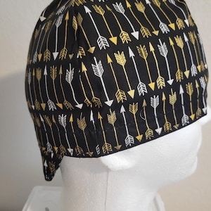 May include: Black cap with a repeating pattern of gold and white arrows. The arrows are arranged in rows, pointing in the same direction. The cap is made of a soft fabric and is designed to fit snugly on the head.
