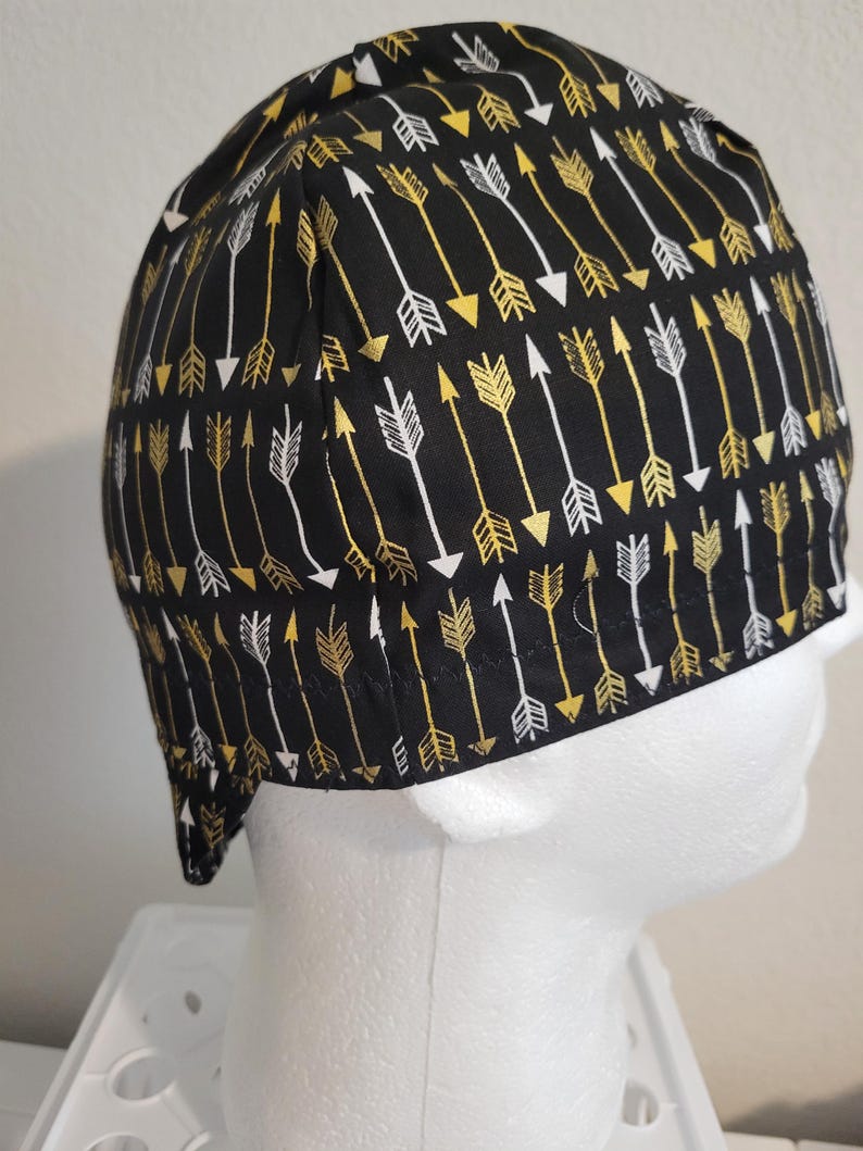 May include: Black cap with a repeating pattern of gold and white arrows. The arrows are arranged in rows, pointing in the same direction. The cap is made of a soft fabric and is designed to fit snugly on the head.