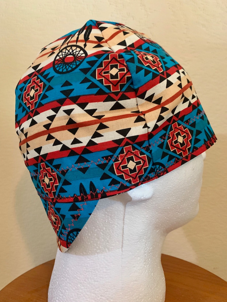 May include: A patterned cap featuring a Native American-inspired design. The cap showcases a vibrant mix of colors, including turquoise, red, black, and beige, with geometric and dreamcatcher motifs. The cap is designed to fit snugly on the head.
