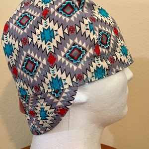 May include: A gray, patterned welding cap featuring a geometric design in teal, red, and white. The cap is made of fabric and has a curved brim. The pattern includes diamond and zigzag shapes.