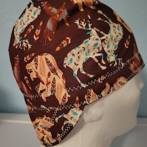 May include: A brown fabric cap featuring a repeating pattern of deer, bears, and feathers in cream, teal, and orange. The cap has a white zigzag stitch along the edge and is designed to fit the head.