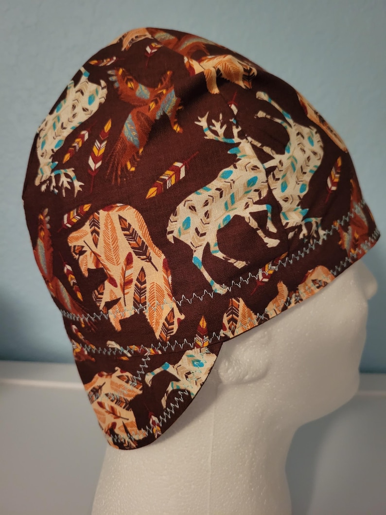 May include: A brown fabric cap featuring a repeating pattern of deer, bears, and feathers in cream, teal, and orange. The cap has a white zigzag stitch along the edge and is designed to fit the head.