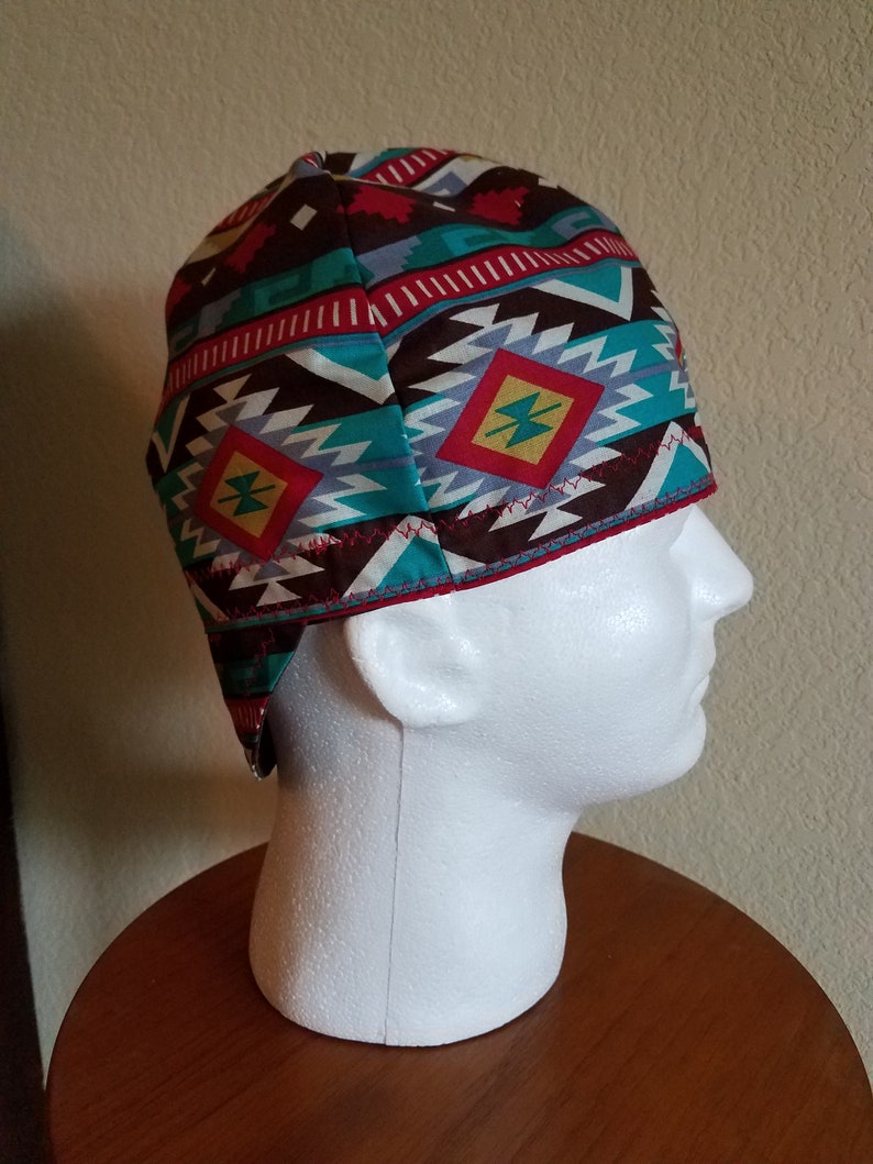 May include: A patterned cap with a Southwestern design. The cap features a repeating geometric pattern in shades of brown, teal, red, and gray. The cap is worn on a white mannequin head, showcasing the design and fit.