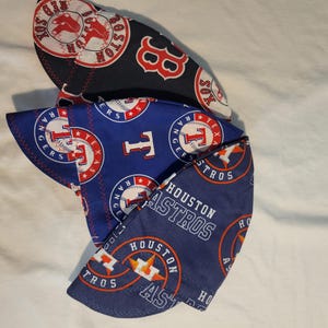 May include: A stack of three baseball-themed face masks. The top mask is black with a red Boston Red Sox logo. The middle mask is blue with a Texas Rangers logo. The bottom mask is blue with a Houston Astros logo.