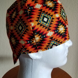 May include: A patterned cap featuring a geometric design in orange, black, white, and teal. The cap is made of fabric and is displayed on a white mannequin head. The cap's design has a Southwestern or Native American aesthetic.