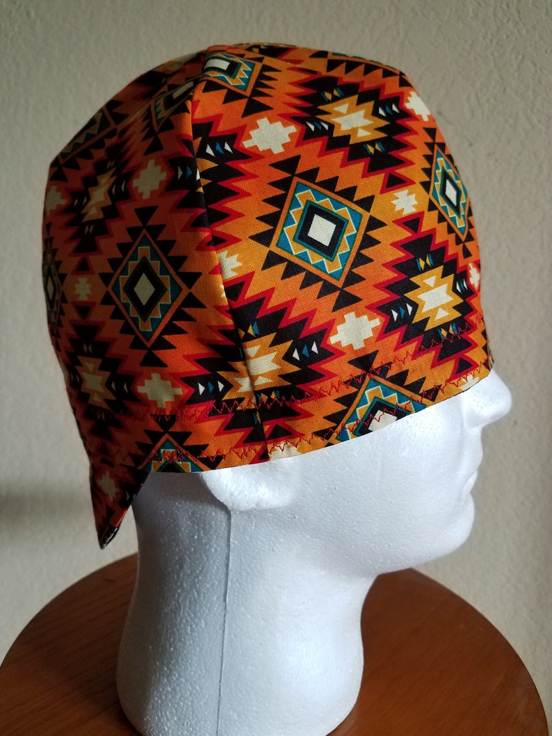 May include: A patterned cap featuring a geometric design in orange, black, white, and teal. The cap is made of fabric and is displayed on a white mannequin head. The cap's design has a Southwestern or Native American aesthetic.