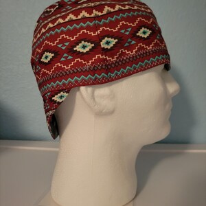 May include: A red and turquoise patterned welding cap. The cap features a repeating geometric design in turquoise, white, and brown on a red background. The cap is displayed on a white mannequin head.