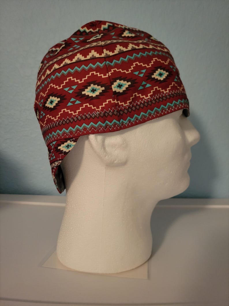 May include: A red and turquoise patterned welding cap. The cap features a repeating geometric design in turquoise, white, and brown on a red background. The cap is displayed on a white mannequin head.