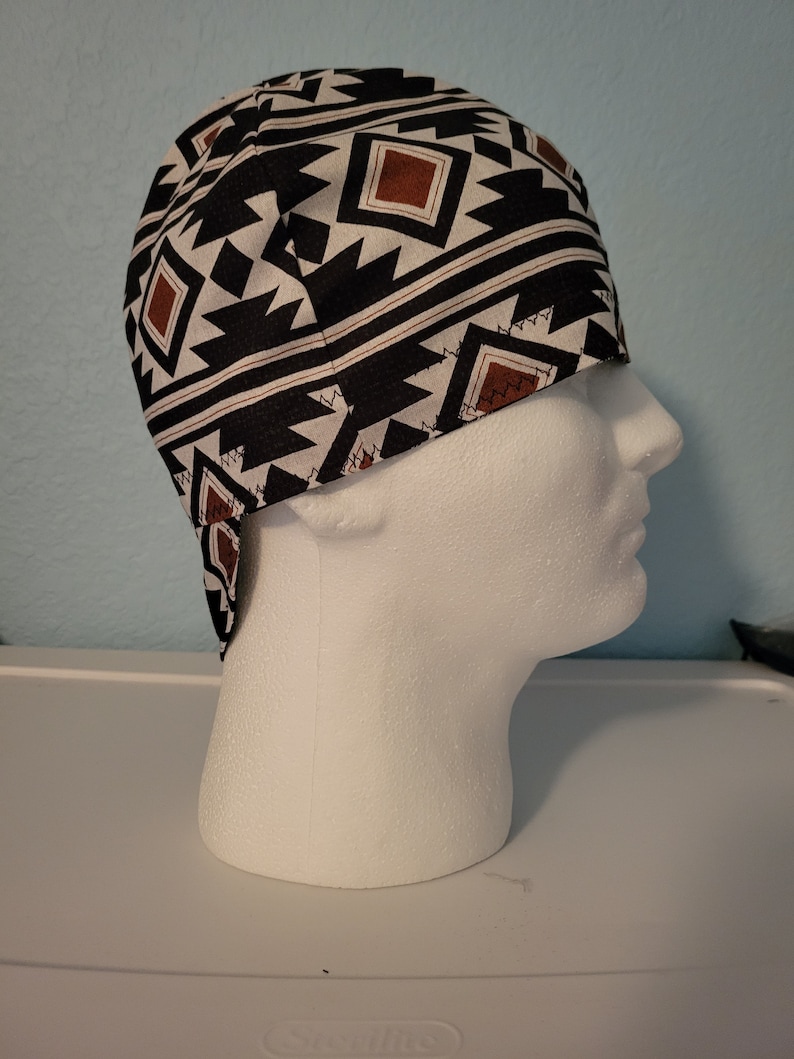 May include: A black, white, and brown patterned welding cap. The cap features a geometric design with brown squares and zig-zag shapes. The cap is made of fabric and is designed to protect the head and hair.