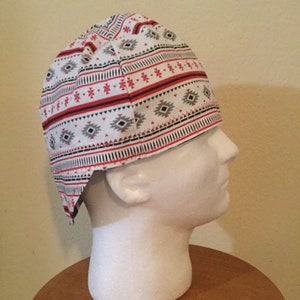 May include: A white head wrap with a geometric pattern in red, black, and gray. The fabric features horizontal stripes and diamond shapes, creating a Southwestern or Native American inspired design. The head covering is displayed on a white mannequin head.