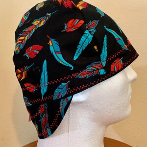 May include: Black welding cap with a colorful feather pattern in shades of red, turquoise, and yellow. The cap has a red zig-zag stitched trim. The cap is designed to fit over the head and ears.