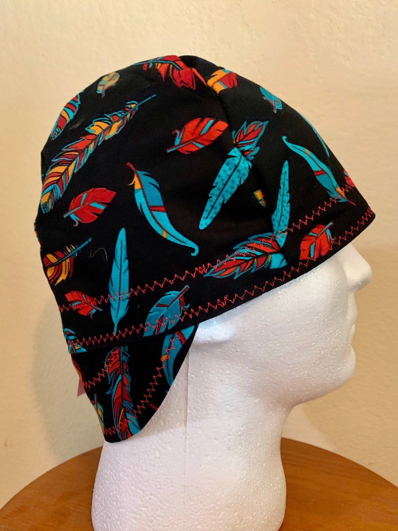 May include: Black welding cap with a colorful feather pattern in shades of red, turquoise, and yellow. The cap has a red zig-zag stitched trim. The cap is designed to fit over the head and ears.