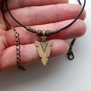 May include: A black leather cord necklace with a bronze arrowhead pendant. The arrowhead has a detailed design and is attached to the cord with small bronze beads.