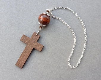 May include: A wooden cross pendant attached to a silver-toned metal chain. The cross features a dark brown wood grain finish. Above the cross, a single round, polished reddish-brown stone bead is accented with decorative silver-toned metal caps.