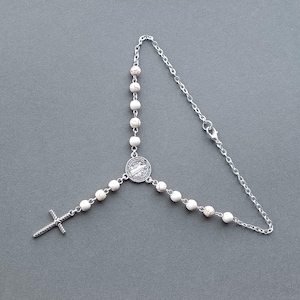Puede incluir: Un rosario de cadena de plata con cuentas blancas y un colgante de plata con una figura religiosa. El rosario tiene una cruz de plata en el extremo.