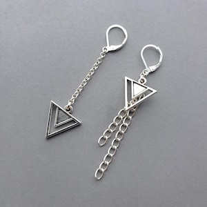 Op de afbeelding: Een paar zilverkleurige hangende oorbellen met geometrische driehoekige bedels. Eén oorbel heeft een enkele ketting die de sluiting met een driehoek verbindt, terwijl de andere een driehoekige bedel heeft met twee bungelende zilveren kettingen.
