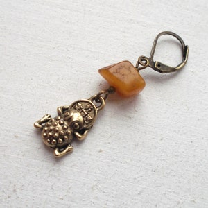 Pode incluir: Brinco dourado com um pequeno amuleto de sapo e uma pedra de cor âmbar marrom. O brinco tem um fecho de gancho.