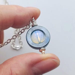 Può includere: Un ciondolo per collana con un anello circolare in ematite grigio scuro che incornicia un cabochon traslucido e iridescente simile all'opale. Il ciondolo è fissato a una catena in metallo argentato, con una piccola perlina argentata alla base.