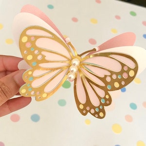 Puede incluir: Una mariposa rosa y dorada con un adorno de perla. La mariposa est&aacute; hecha de papel y tiene un efecto 3D.