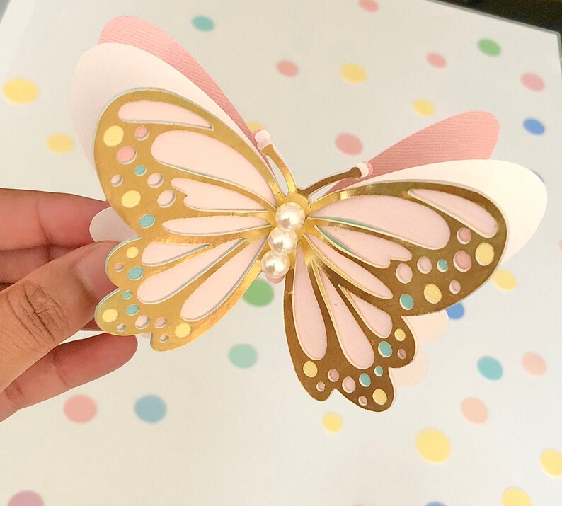 Puede incluir: Una mariposa rosa y dorada con un adorno de perla. La mariposa est&aacute; hecha de papel y tiene un efecto 3D.