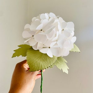 Op de afbeelding: Een handgemaakte witte papieren hortensia bloem met groene papieren bladeren en een groene stengel. De bloemblaadjes zijn gelaagd om een volle bloei te creëren. De bladeren hebben een gekartelde rand.