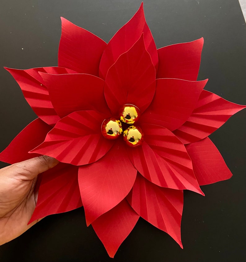 May include: A red paper poinsettia flower with three gold ornaments in the center.