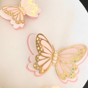 Puede incluir: Dos mariposas de capas rosa y doradas con detalles de perlas sobre un fondo blanco.