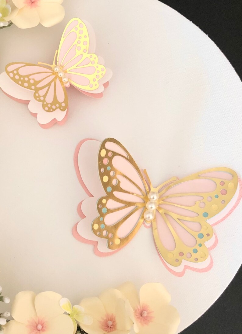 Puede incluir: Dos mariposas de capas rosa y doradas con detalles de perlas sobre un fondo blanco.