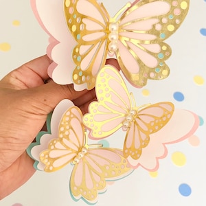 Puede incluir: Tres mariposas de papel rosa y dorado con detalles de perlas. Las mariposas est&aacute;n superpuestas y tienen un dise&ntilde;o delicado e intrincado.
