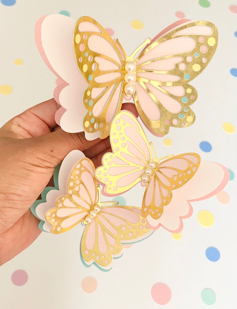 Puede incluir: Tres mariposas de papel rosa y dorado con detalles de perlas. Las mariposas est&aacute;n superpuestas y tienen un dise&ntilde;o delicado e intrincado.
