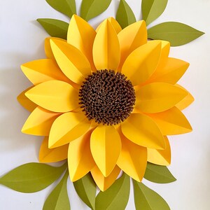 May include: A large paper sunflower with yellow petals and a brown center. The sunflower is surrounded by green leaves.