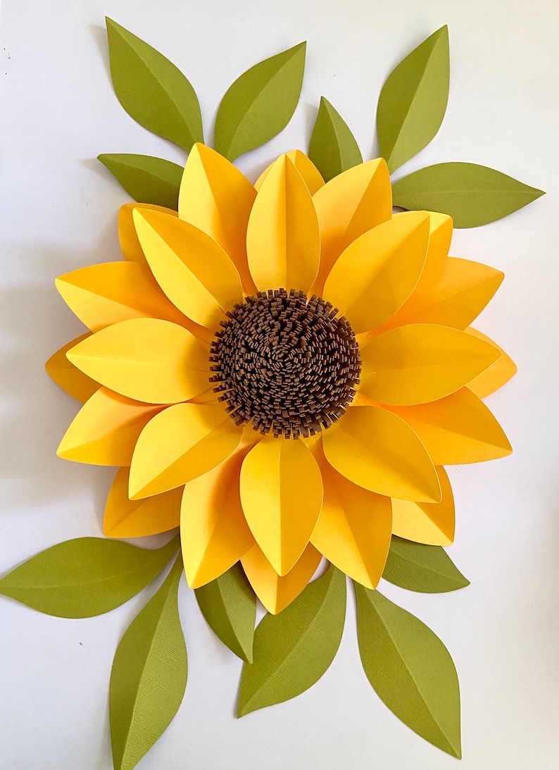 May include: A large paper sunflower with yellow petals and a brown center. The sunflower is surrounded by green leaves.