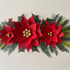May include: A paper craft Christmas garland with three red poinsettia flowers, green holly leaves, and gold glitter balls.