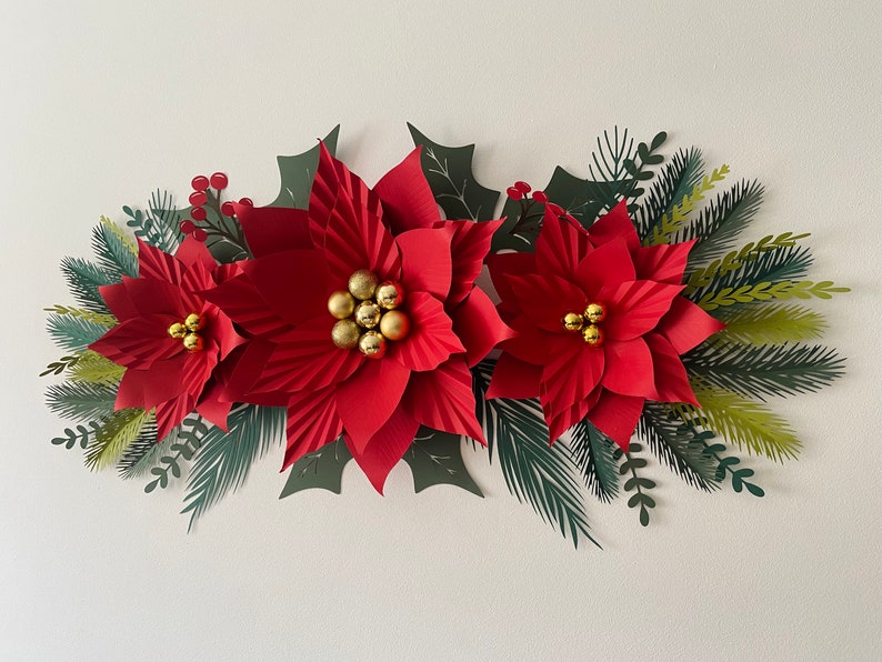 May include: A paper craft Christmas garland with three red poinsettia flowers, green holly leaves, and gold glitter balls.