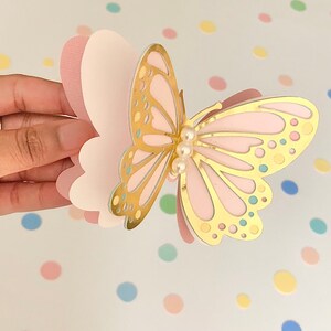 Puede incluir: Una manualidad de papel de mariposa rosa y dorada con un adorno de perla. La mariposa est&aacute; hecha de capas de papel rosa y tiene un contorno dorado.