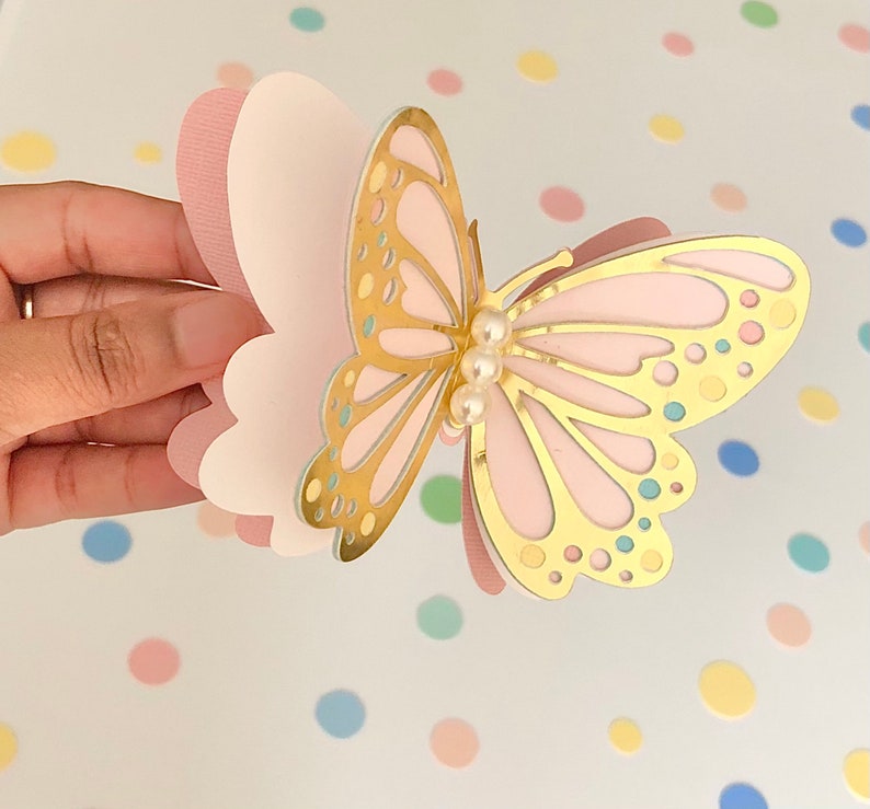 Puede incluir: Una manualidad de papel de mariposa rosa y dorada con un adorno de perla. La mariposa est&aacute; hecha de capas de papel rosa y tiene un contorno dorado.