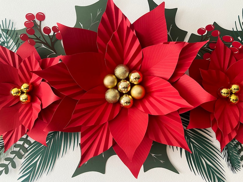 May include: Three large red paper poinsettia flowers with gold glitter ornaments in the center. The flowers are surrounded by green paper leaves.