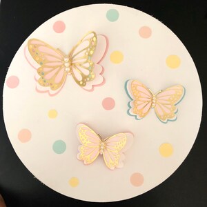 Puede incluir: Tres mariposas de papel con detalles dorados y centros de perlas sobre un fondo blanco con lunares de color rosa pastel, amarillo y azul.