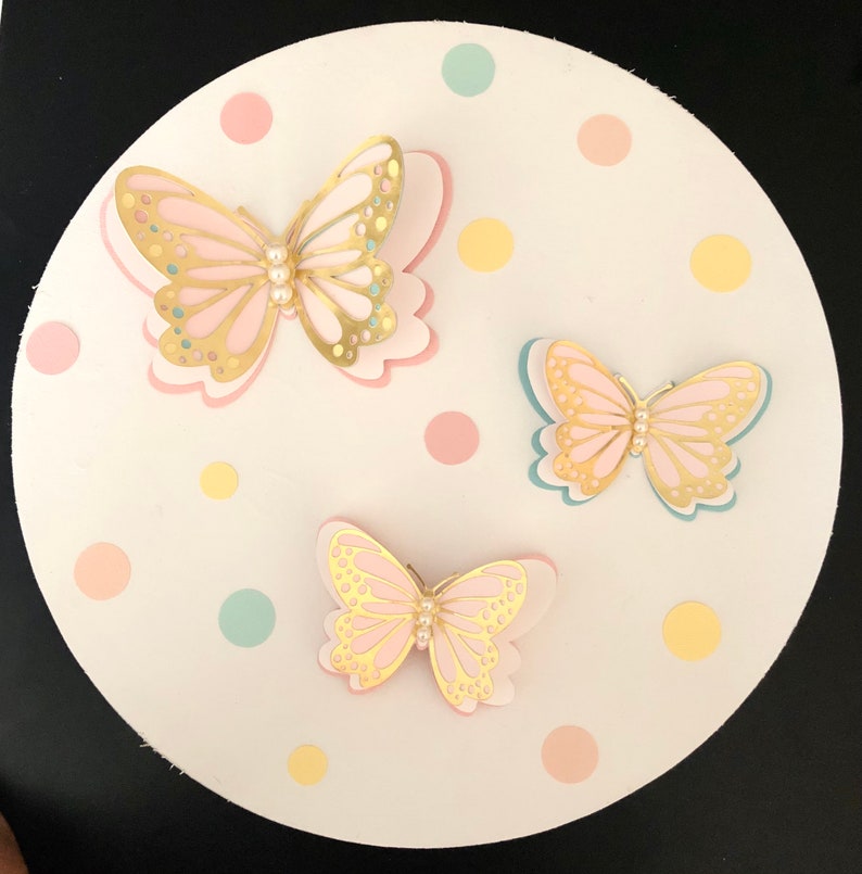 Puede incluir: Tres mariposas de papel con detalles dorados y centros de perlas sobre un fondo blanco con lunares de color rosa pastel, amarillo y azul.