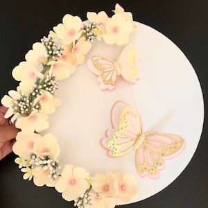 Puede incluir: Un tablero circular blanco para pastel decorado con una corona de flores de papel amarillo p&aacute;lido y dos mariposas rosadas con detalles dorados. Las mariposas tienen adornos de perlas.