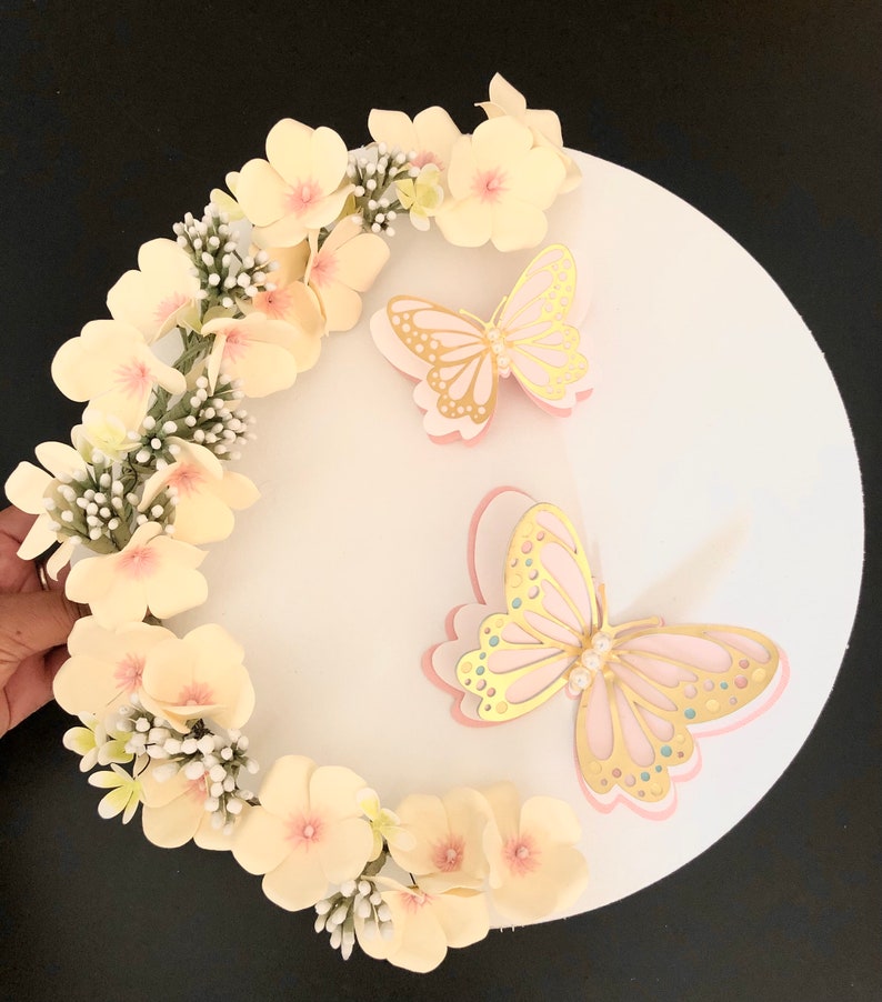 Puede incluir: Un tablero circular blanco para pastel decorado con una corona de flores de papel amarillo p&aacute;lido y dos mariposas rosadas con detalles dorados. Las mariposas tienen adornos de perlas.