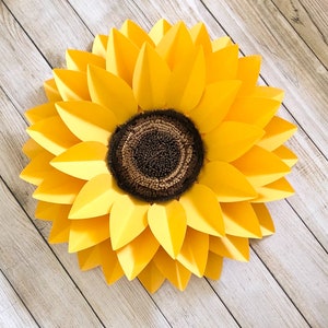 May include: A large paper sunflower with yellow petals and a brown center. The sunflower is on a light wood background.