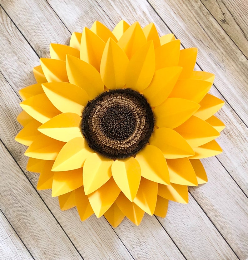May include: A large paper sunflower with yellow petals and a brown center. The sunflower is on a light wood background.