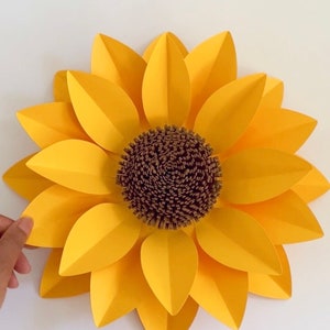 May include: A large paper sunflower with yellow petals and a brown center. The sunflower is made of paper and has a 3D effect.