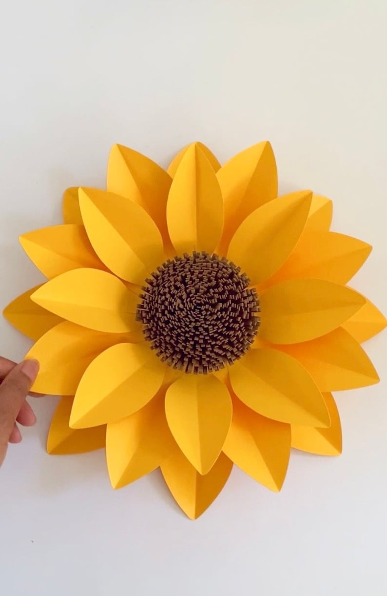 May include: A large paper sunflower with yellow petals and a brown center. The sunflower is made of paper and has a 3D effect.