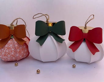 Puede incluir: Tres adornos decorativos de papel con lazos. Uno es naranja con un estampado floral, otro es blanco con un lazo verde y el tercero es blanco con un lazo rojo. Hay cuentas doradas dispersas alrededor de los adornos.