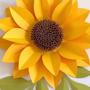 May include: A large paper sunflower with yellow petals and a brown center. The sunflower is surrounded by green paper leaves.